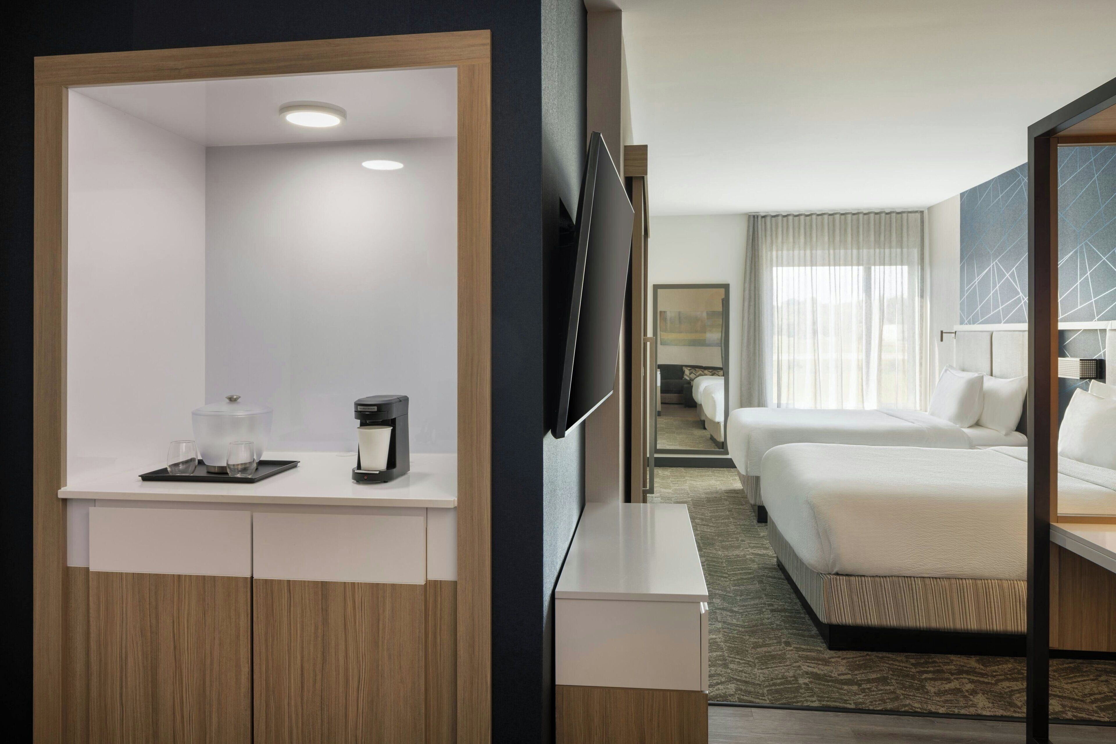 Habitación de hotel con cama, televisor, cafetera y una pequeña mesa con vasos y una taza.