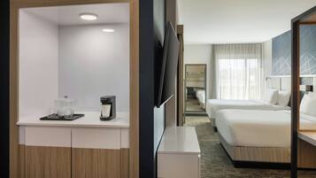 Habitación de hotel con cama, televisor, cafetera y una pequeña mesa con vasos y una taza.