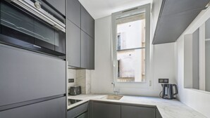 A modern kitchen with dark cabinetry, a marble countertop, a built-in toaster, and a window providing natural light. - Terne Maillot - Gaston Bertandeau 1dbr (Paris)