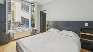 A bedroom with a bed, a window with an open door, a radiator, and a view of a building outside. - Terne Maillot - Gaston Bertandeau 1dbr (Paris)