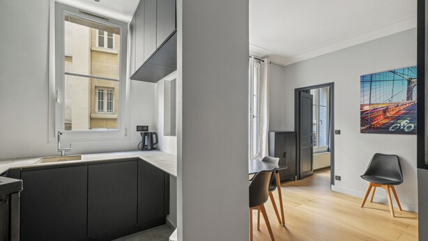 A modern kitchen with a large island, a dining area with two chairs, and a view of the city through a window. - Terne Maillot - Gaston Bertandeau 1dbr (Paris)
