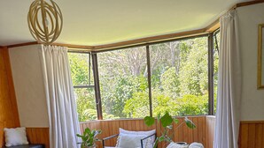 Un salon moderne avec un grand canapé, une table basse en bois et un fauteuil. On y trouve une fenêtre avec des rideaux, une plante et une vue sur la verdure à l’extérieur.
