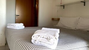 A hotel room with a checkered bedspread, a neatly folded white towel on the bed, a wooden door, and a white wall with a light switch and a red cord. - Palme 8 - Marcelli, three-room apartment with private garden, Residence Le Palme (Marcelli)