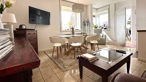 Een moderne woonkamer met een eethoek, een aan de muur bevestigde televisie en een groot raam dat voor natuurlijk licht zorgt.