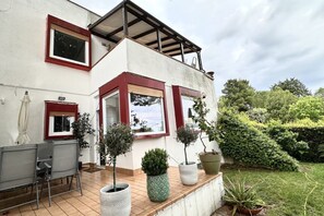 A two-story house with a patio, potted plants, and a view of the surrounding greenery.