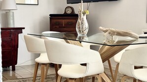 Een eetruimte met een glazen tafel, witte stoelen, een houten kast, een klok en een vaas met bloemen.