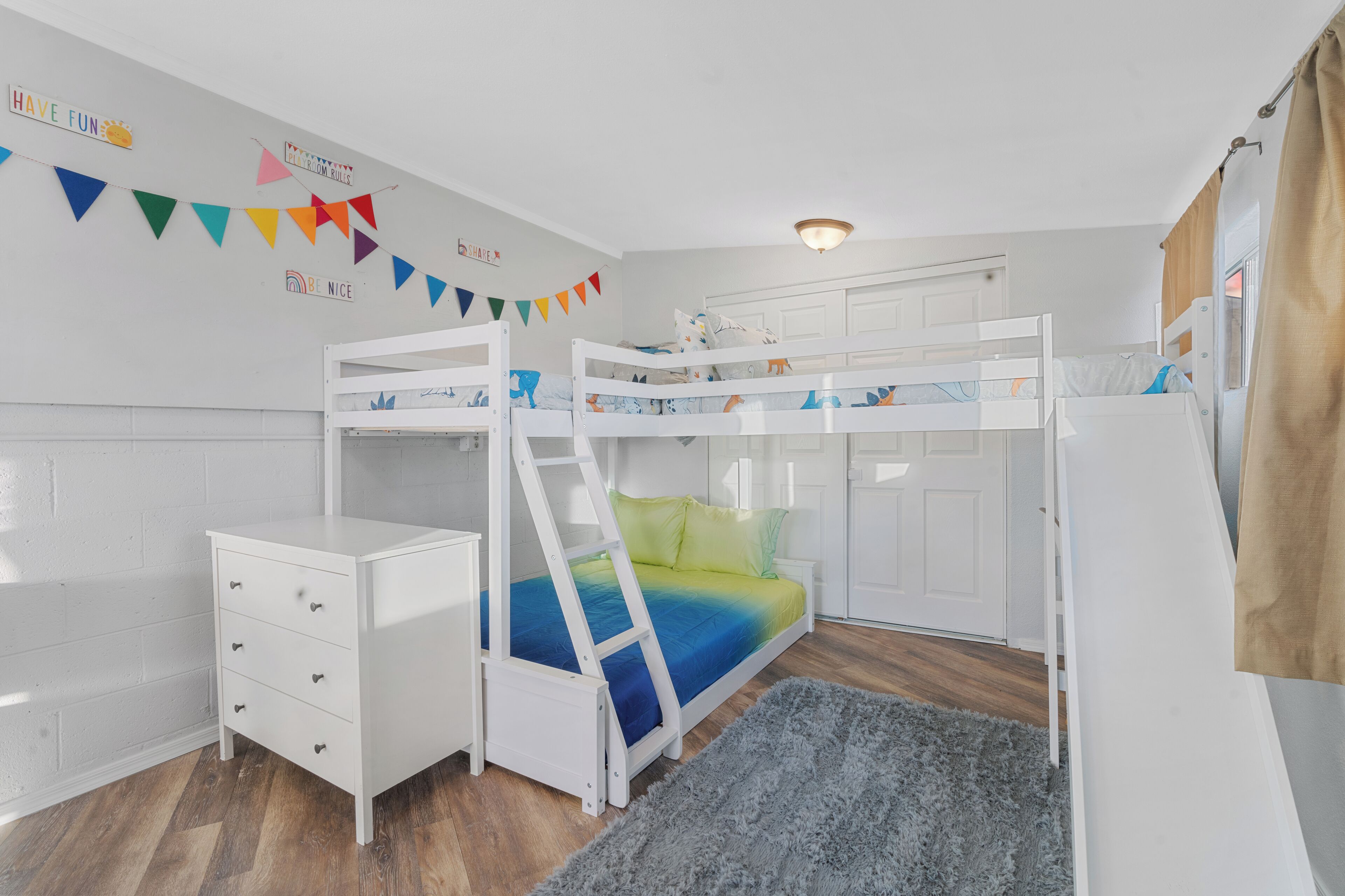 Une chambre d’enfant avec des lits superposés, une commode blanche et une grande banderole colorée accrochée au mur.