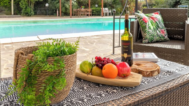 Area piscina con un cesto in vimini, un vassoio di legno con frutta e una bottiglia, e un divano in vimini con un cuscino.