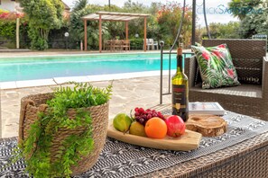 A poolside setting with a wicker basket, a wooden tray with fruits and a bottle, and a wicker sofa with a cushion.