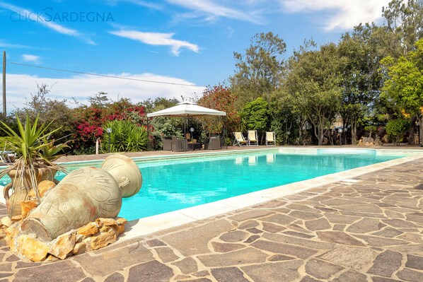A swimming pool with a stone deck, surrounded by trees and a covered seating area.