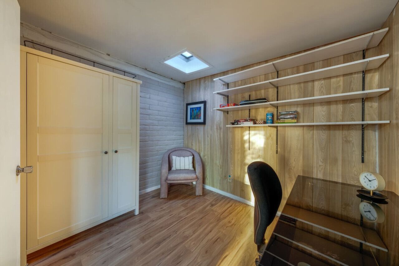 A room with a wooden floor, a white wardrobe, a small chair, a shelf with books and a clock, and a skylight.