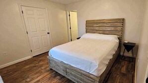 A bedroom with a wooden bed frame, a white bedspread, a bedside table with a lamp, and a door leading to another room. - New listing! Perfectly centered in the Birch Harbor village!

 (Gouldsboro)