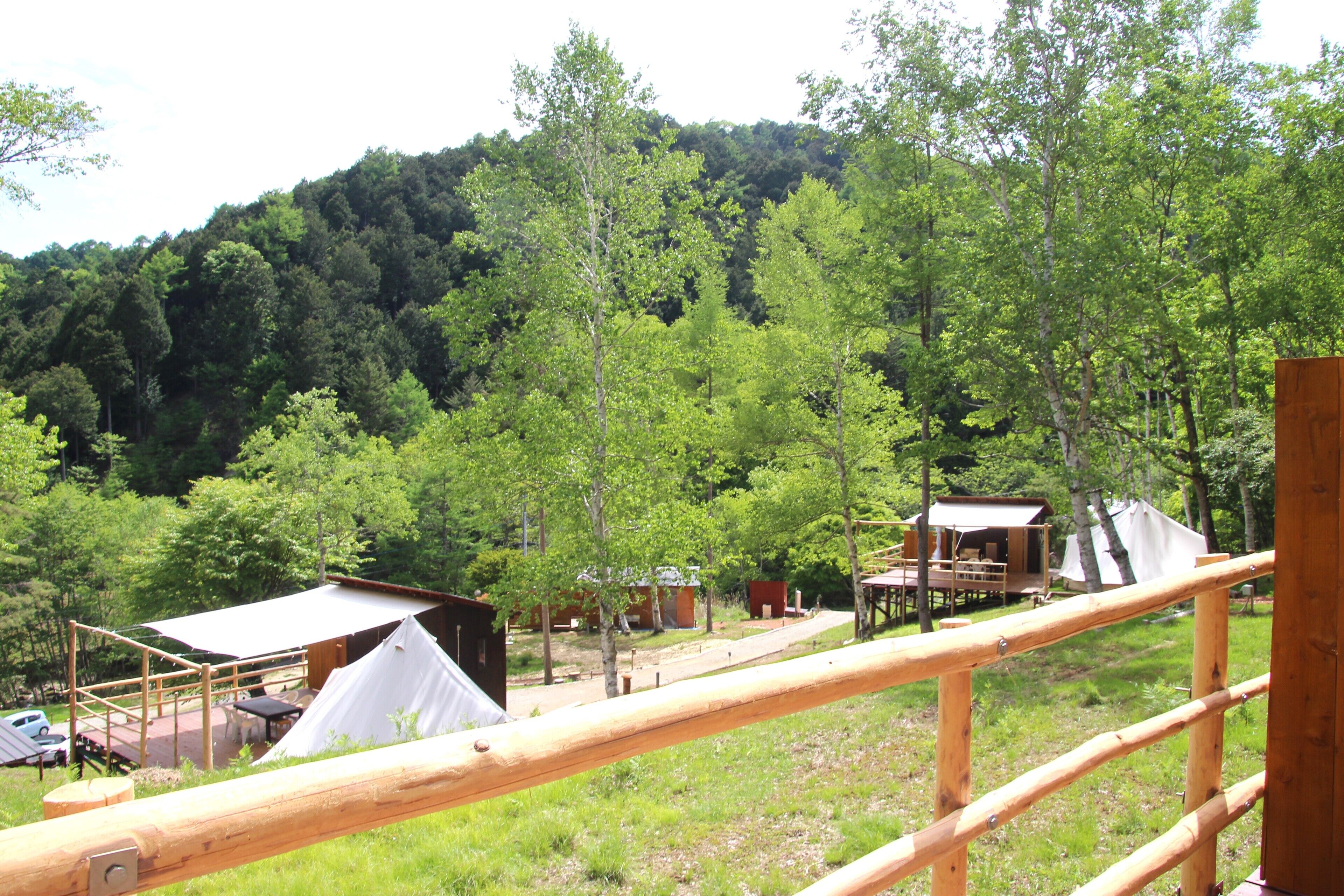 A campsite with wooden structures, including a tent and a cabin, surrounded by trees and a hill in the background.