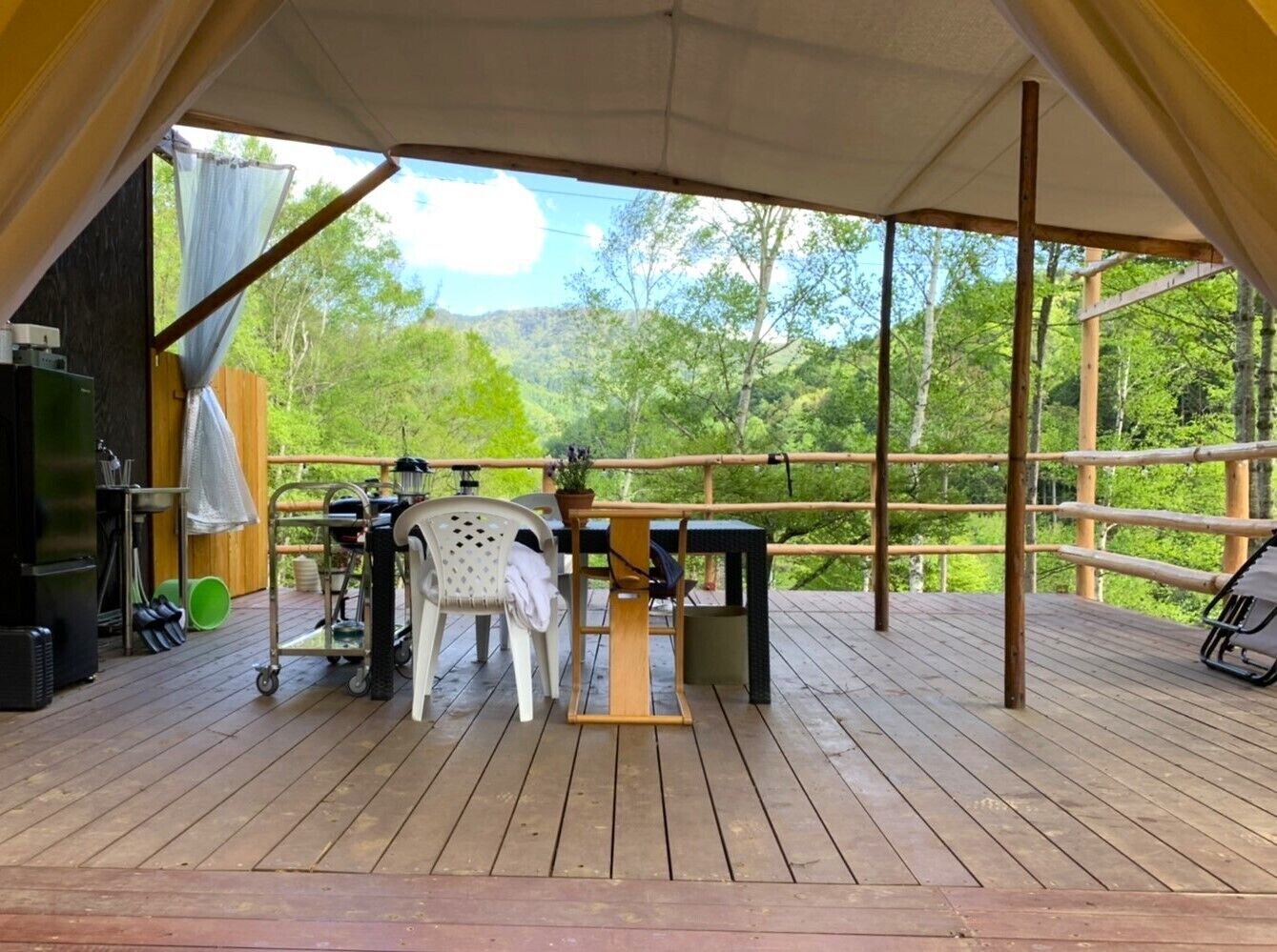 En udendørs træterrasse med et bord og stole, omgivet af et træhegn, med udsigt over et skovområde.