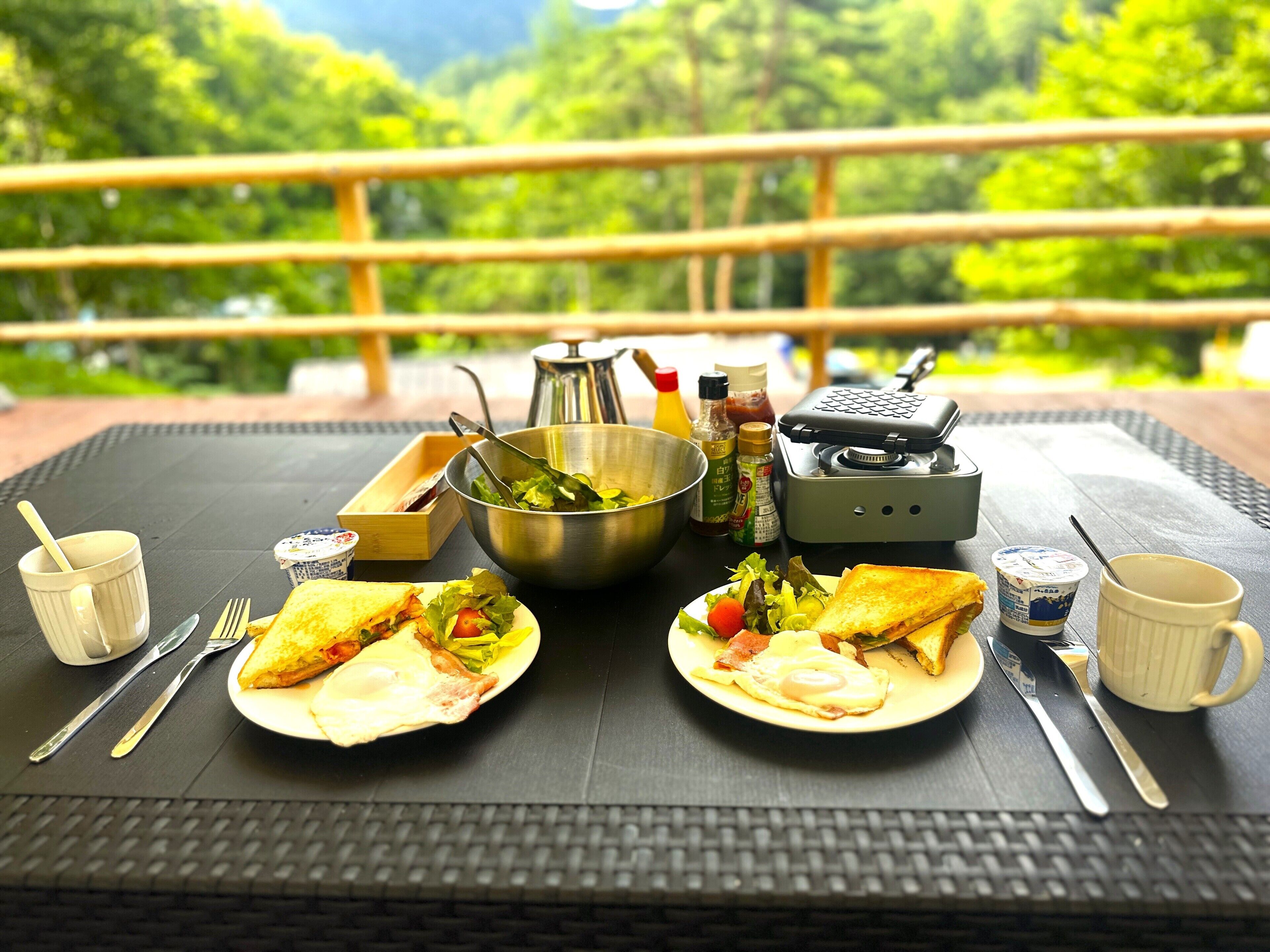 Mesa de comedor con comida, utensilios, condimentos y una estufa portátil, ubicada al aire libre con vista a la vegetación.