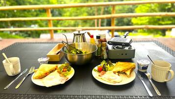 Mesa de comedor con comida, utensilios, condimentos y una estufa portátil, ubicada al aire libre con vista a la vegetación.