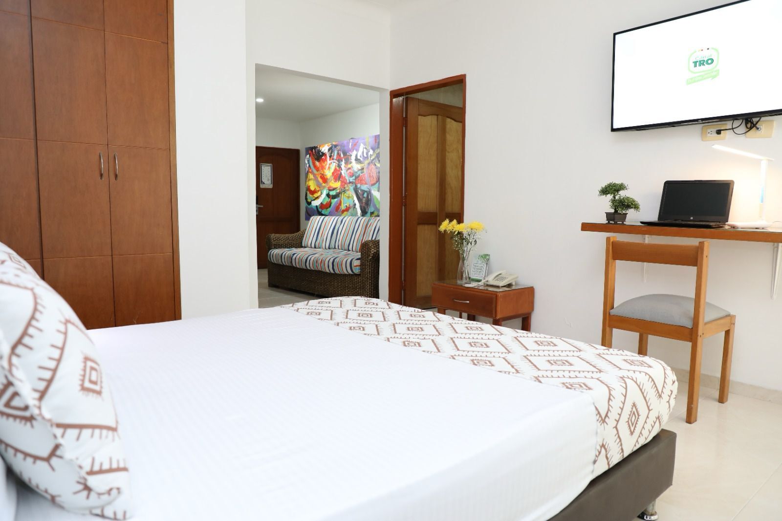 A hotel room with a bed, a desk with a laptop, a chair, a wardrobe, and a colorful painting on the wall.