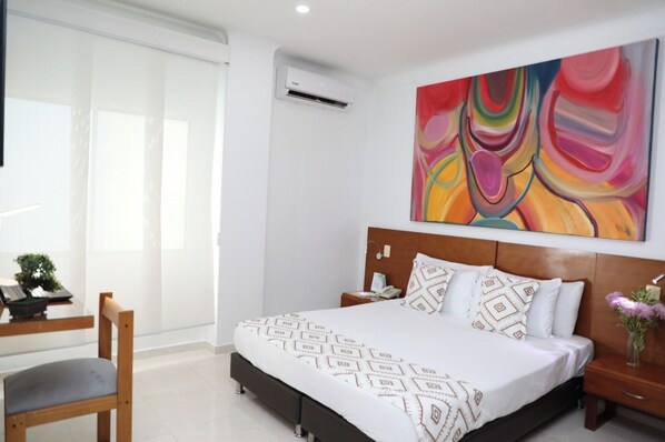 A modern hotel room with a large bed, a desk with a chair, a window with blinds, and a colorful abstract painting on the wall. - Hotel Guane (Bucaramanga)