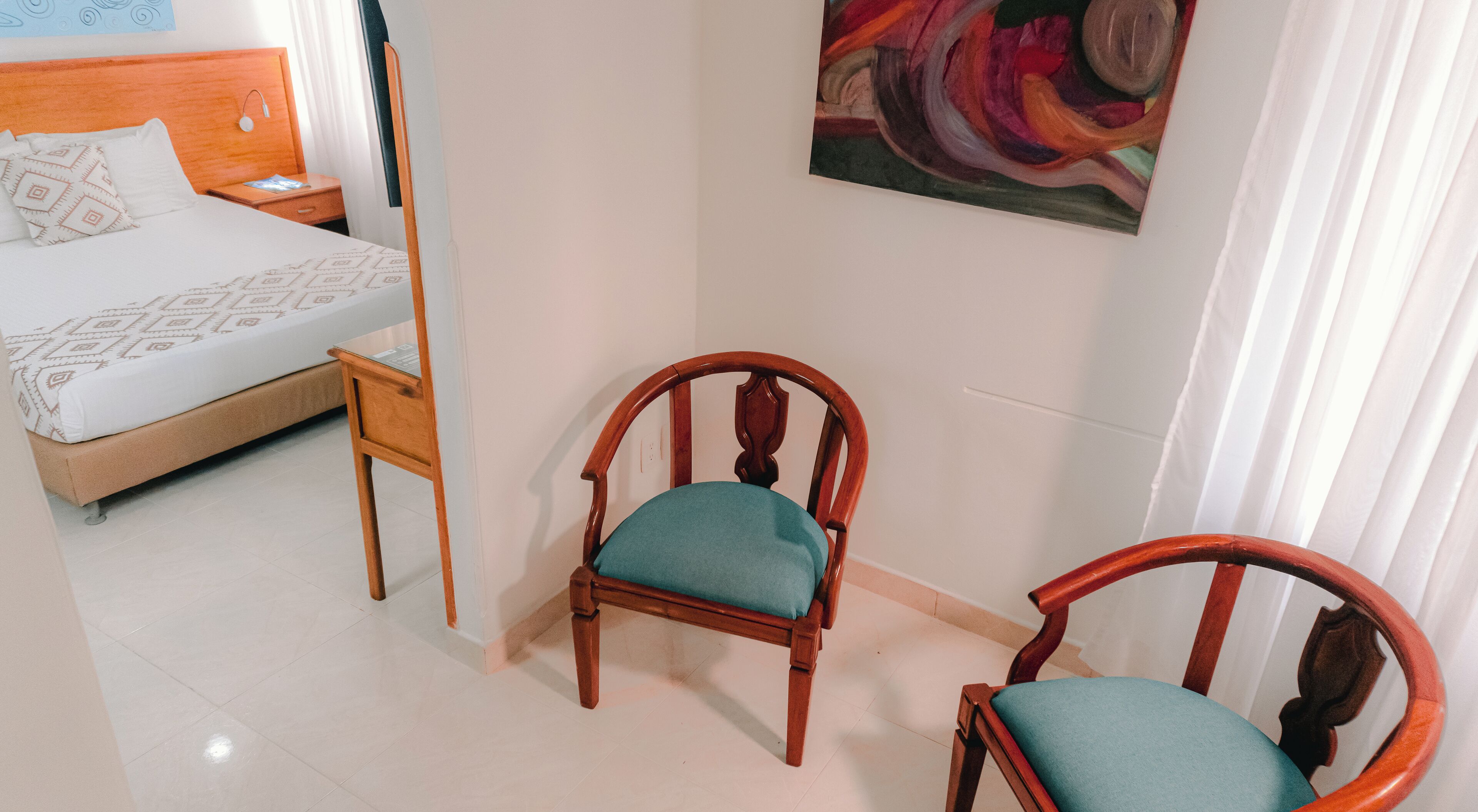 Una habitación de hotel con una cama, una silla de madera con cojín color aguamarina y un cuadro en la pared.