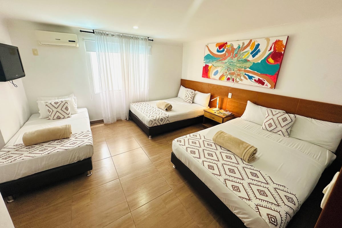 A hotel room with two single beds, a double bed, a TV, an air conditioner, a window with curtains, and a colorful abstract painting on the wall.