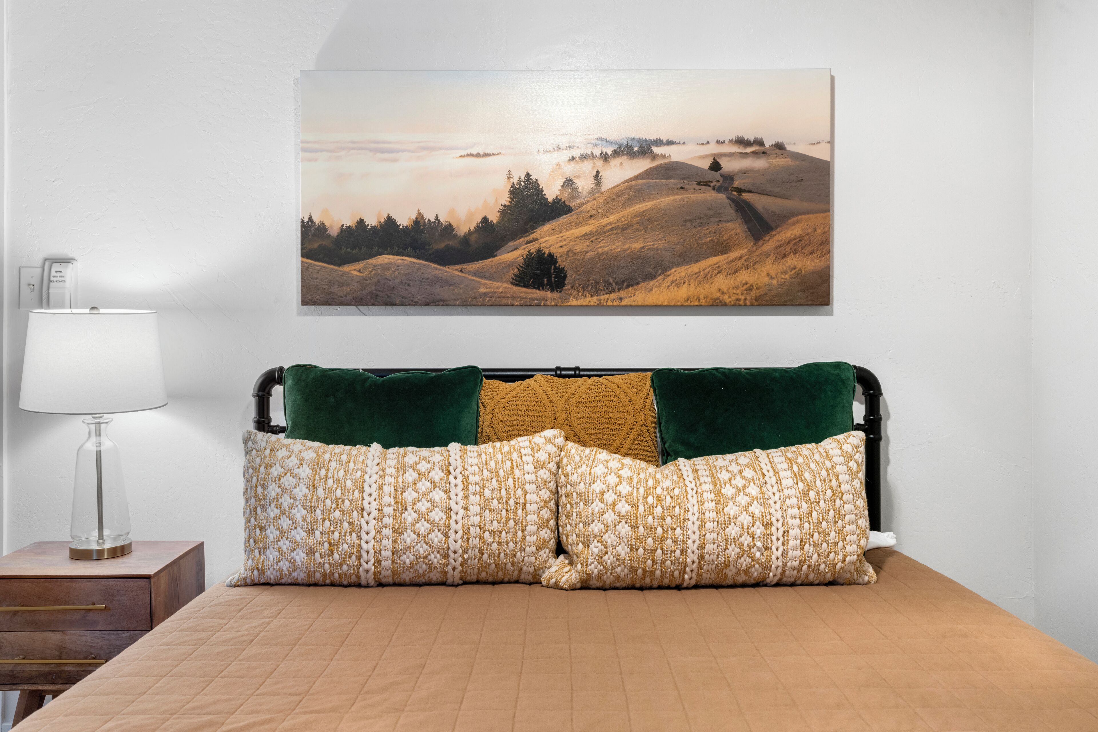 Una camera da letto con un letto, cuscini, un comodino, una lampada e un dipinto che raffigura un paesaggio.