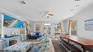 Ein geräumiges Wohnzimmer mit großem Fernseher, Deckenventilator, Essbereich mit Holztisch und Bänken sowie einem blauen Sofa.
