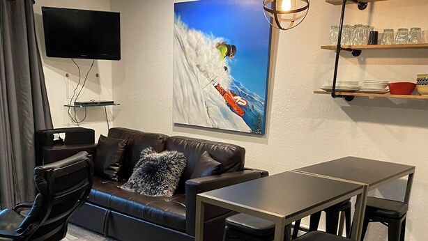 A modern living room with a leather sofa, a flat-screen TV, a dining table with stools, and a painting of a skier on a mountain. - Beautiful Girdwood Condo (Anchorage)