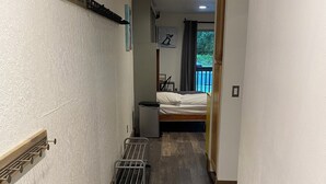 A narrow hallway with a wooden floor, a bed with a white mattress, a small trash bin, and a metal rack on the wall. - Beautiful Girdwood Condo (Anchorage)