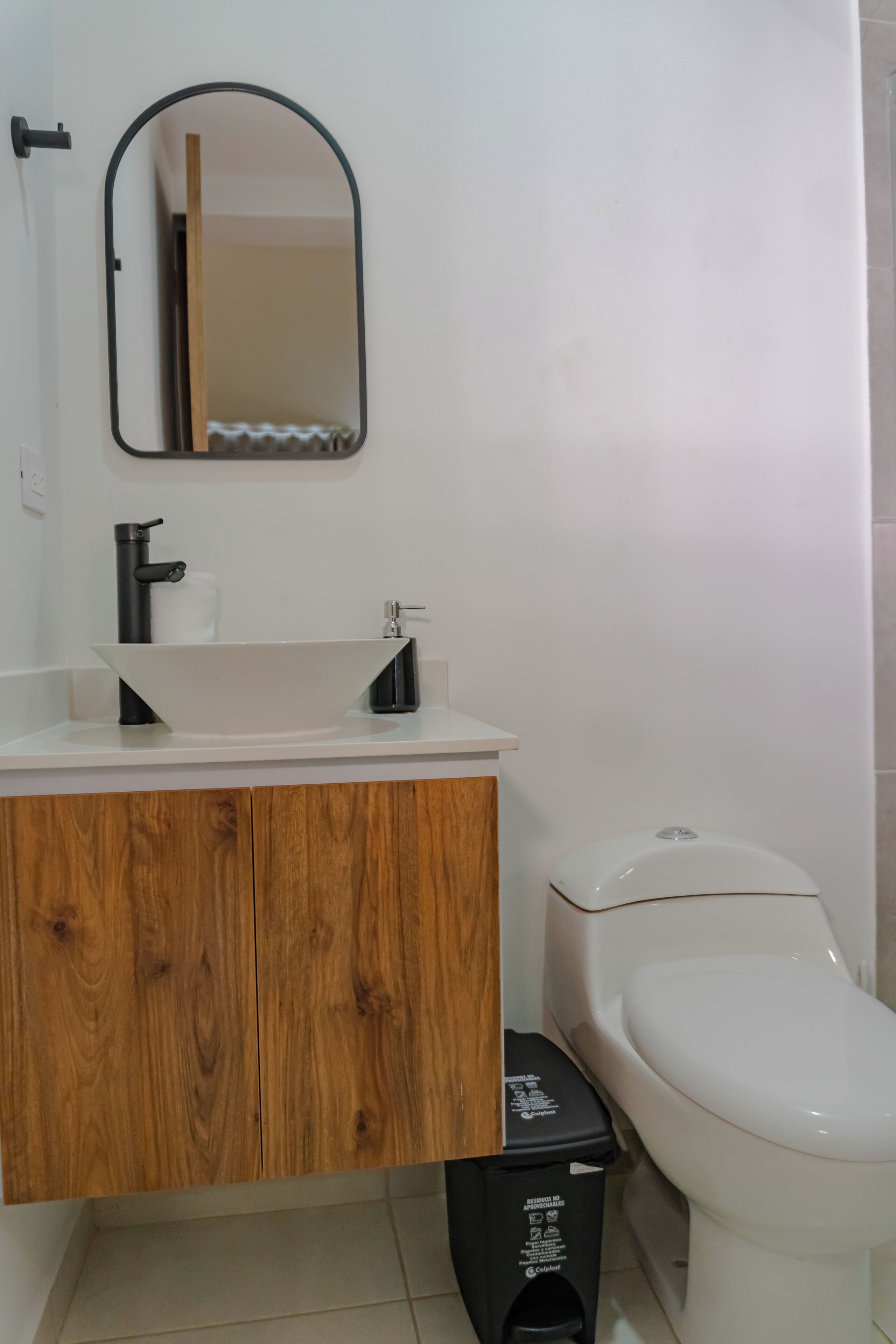A bathroom with a white sink, black faucet, wooden vanity, white toilet, and a black trash bin.