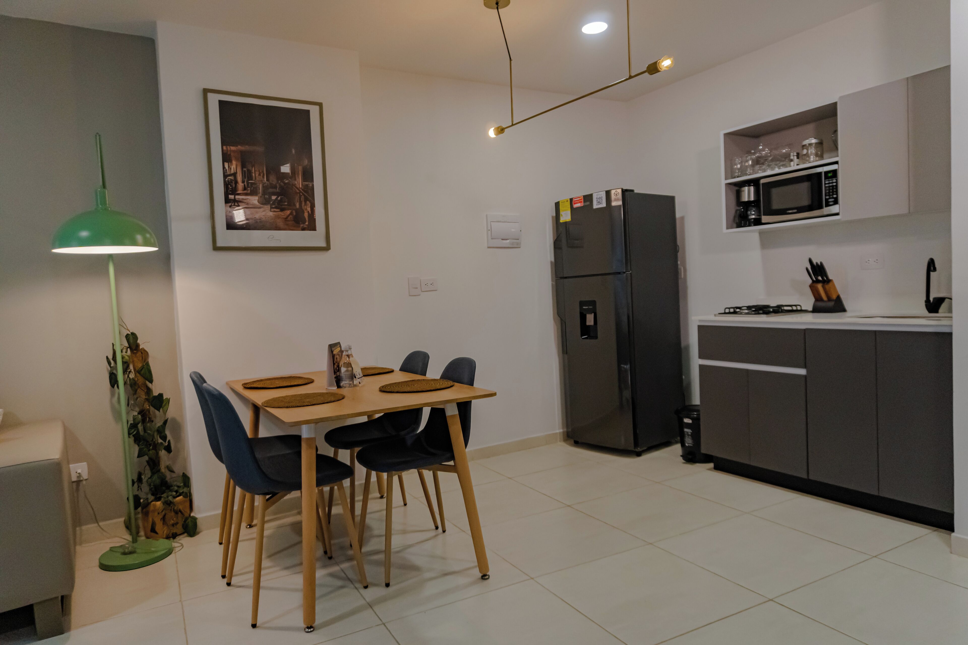 Cocina moderna con comedor, que incluye una mesa con sillas, un refrigerador, un microondas y una lámpara colgante verde.