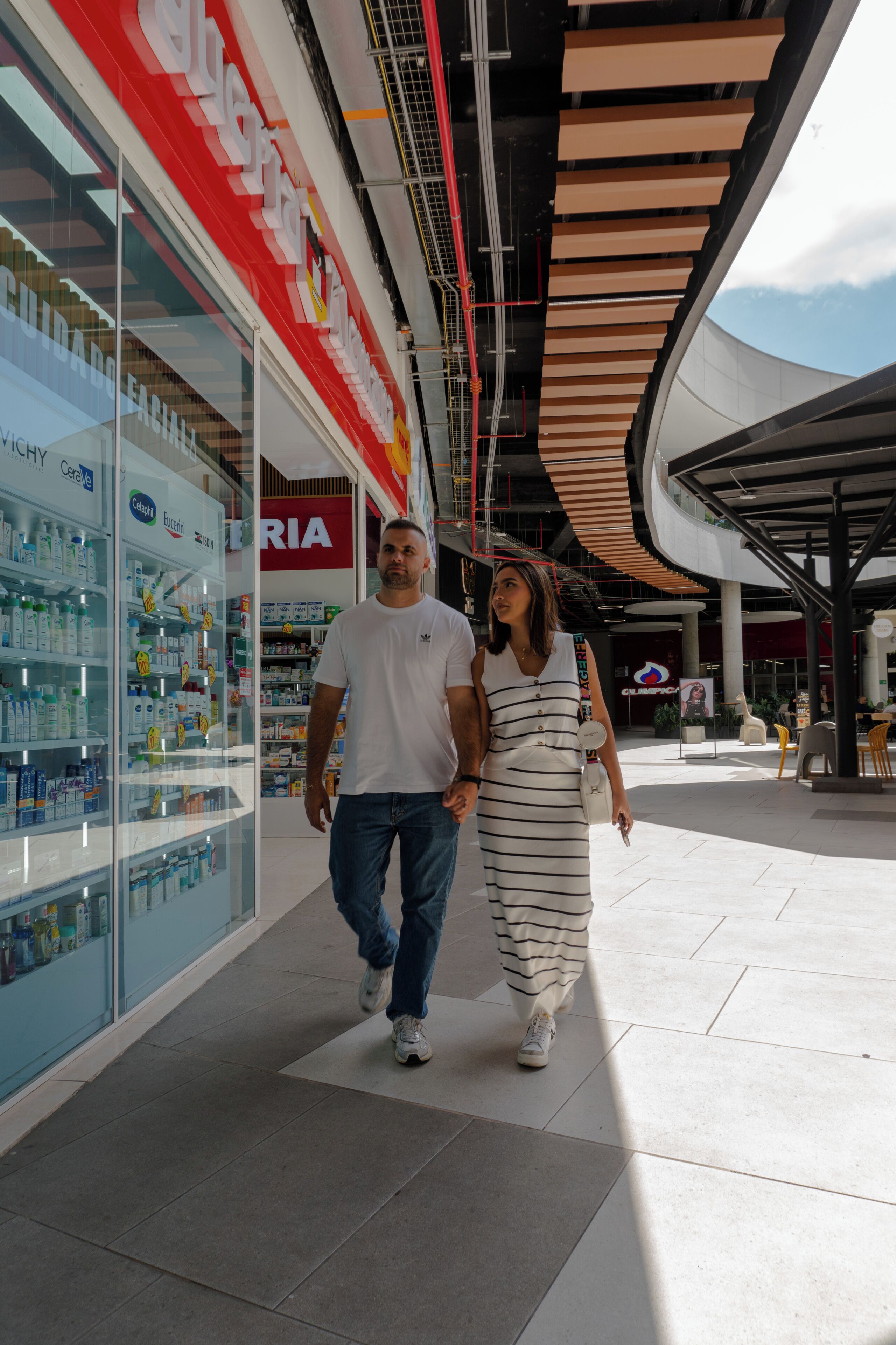 A couple walking in a shopping mall corridor with a pharmacy on the left and a curved ceiling structure on the right.