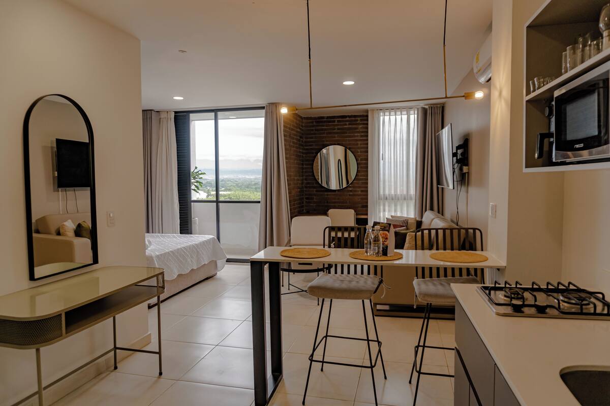 A modern hotel room with a balcony, a dining area, a kitchen, and a TV.