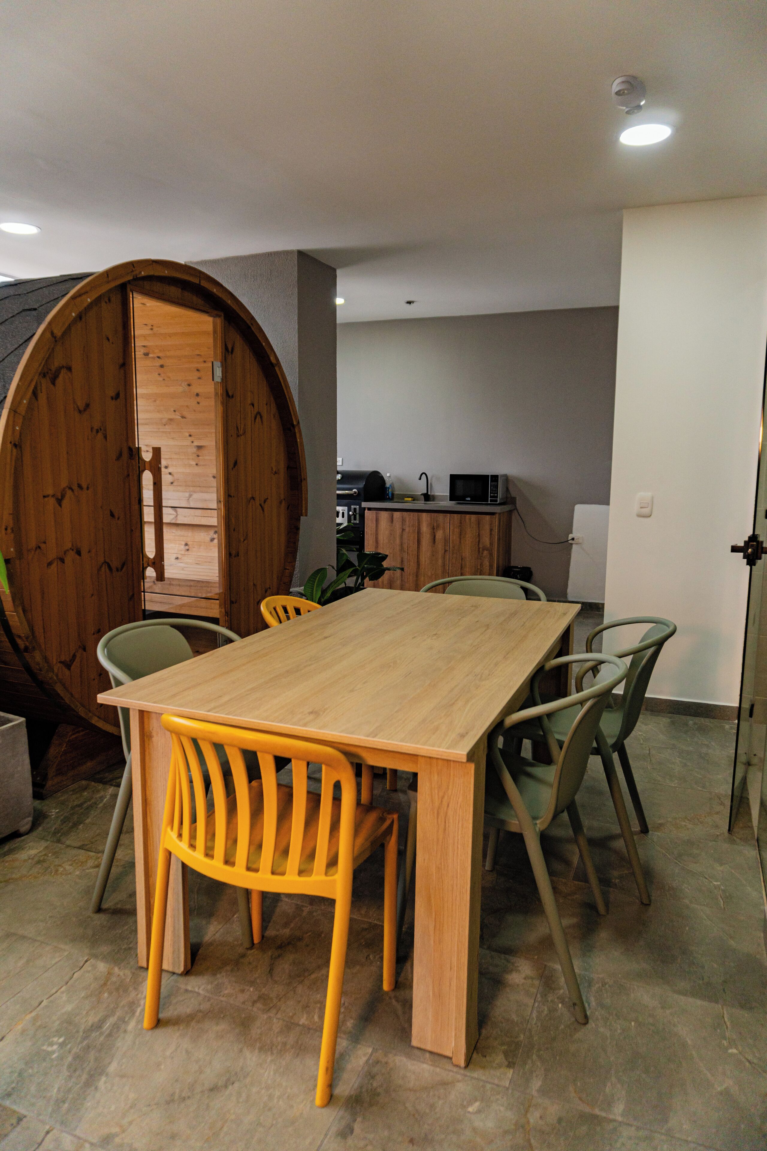 Ein Essbereich mit Holztisch und -stühlen, im Hintergrund eine Küche und eine große Holzfass-Konstruktion.