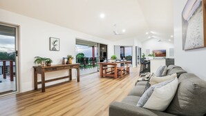 A spacious living area with a wooden floor, a grey sofa, a dining table with chairs, and a balcony with a view.