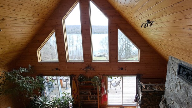 Ein Raum mit hohen Decken, Holzwänden und großen Fenstern, eine Steinwand mit einer Gedenktafel und ein Kamin mit Holzscheiten.