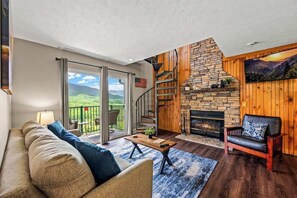 A spacious living room with a fireplace, a wooden staircase, and a balcony with a scenic view.
