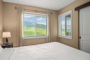 A bedroom with a large bed, two windows with curtains, a nightstand with a lamp, and a view of green hills.