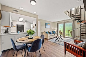 A modern kitchen with white cabinets, a dining area with a round table and blue chairs, a living room with a sofa and a view of the outdoors.