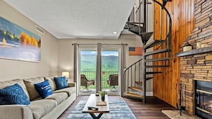 A living room with a sofa, coffee table, and a fireplace. There is a staircase leading to a balcony with a view of the mountains. - Mountain Views | Indoor Pool & Hot Tub Access (Gatlinburg)