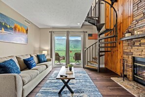 Ein Wohnzimmer mit Sofa, Couchtisch und Kamin. Eine Treppe führt zu einem Balkon mit Blick auf die Berge.