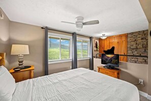 A bedroom with a bed, a nightstand with a lamp, a television, and a view of a mountain through large windows. - Mountain Views | Indoor Pool & Hot Tub Access (Gatlinburg)