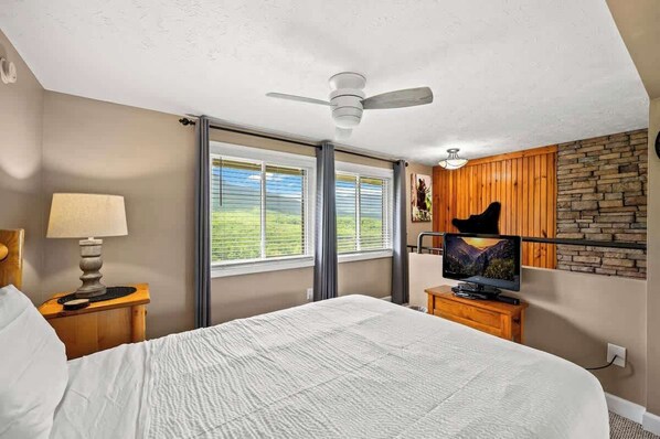 A bedroom with a bed, a nightstand with a lamp, a television, and a view of a mountain through large windows.