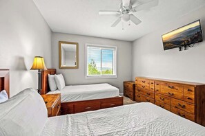 Une chambre avec deux lits, une commode en bois, un ventilateur de plafond, une fenêtre avec des stores et un téléviseur fixé au mur.