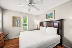 A bedroom with a large bed, a ceiling fan, a window with a view, a bedside lamp, and a framed picture on the wall.