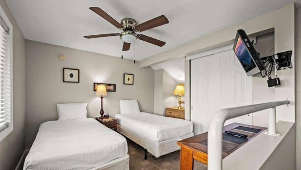 A hotel room with two single beds, a ceiling fan, bedside lamps, a TV mounted on the wall, and a small table with a remote control.
