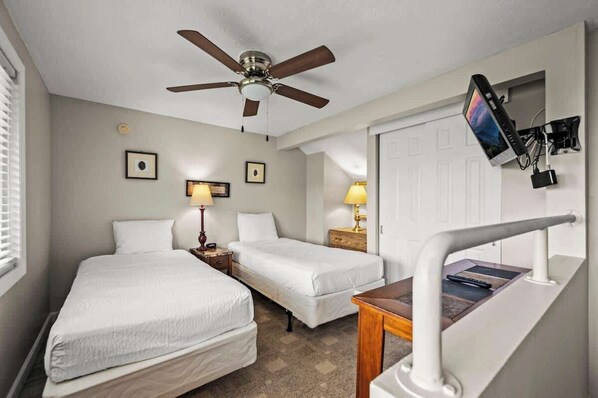 Habitación de hotel con dos camas individuales, un ventilador de techo, lámparas de noche, un televisor montado en la pared y una mesita con el control remoto.