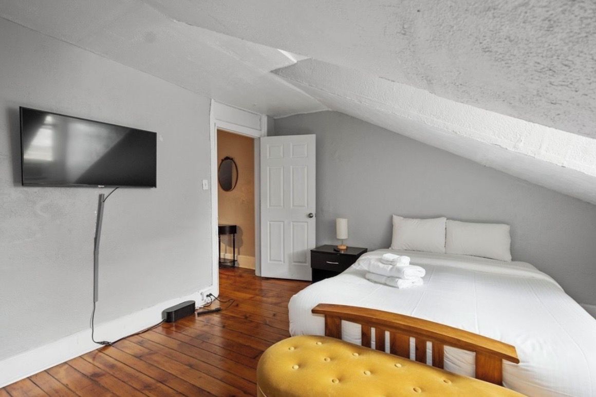 A bedroom with a flat-screen TV mounted on the wall, a bed with white linens, a wooden bench, and a door leading to another room.