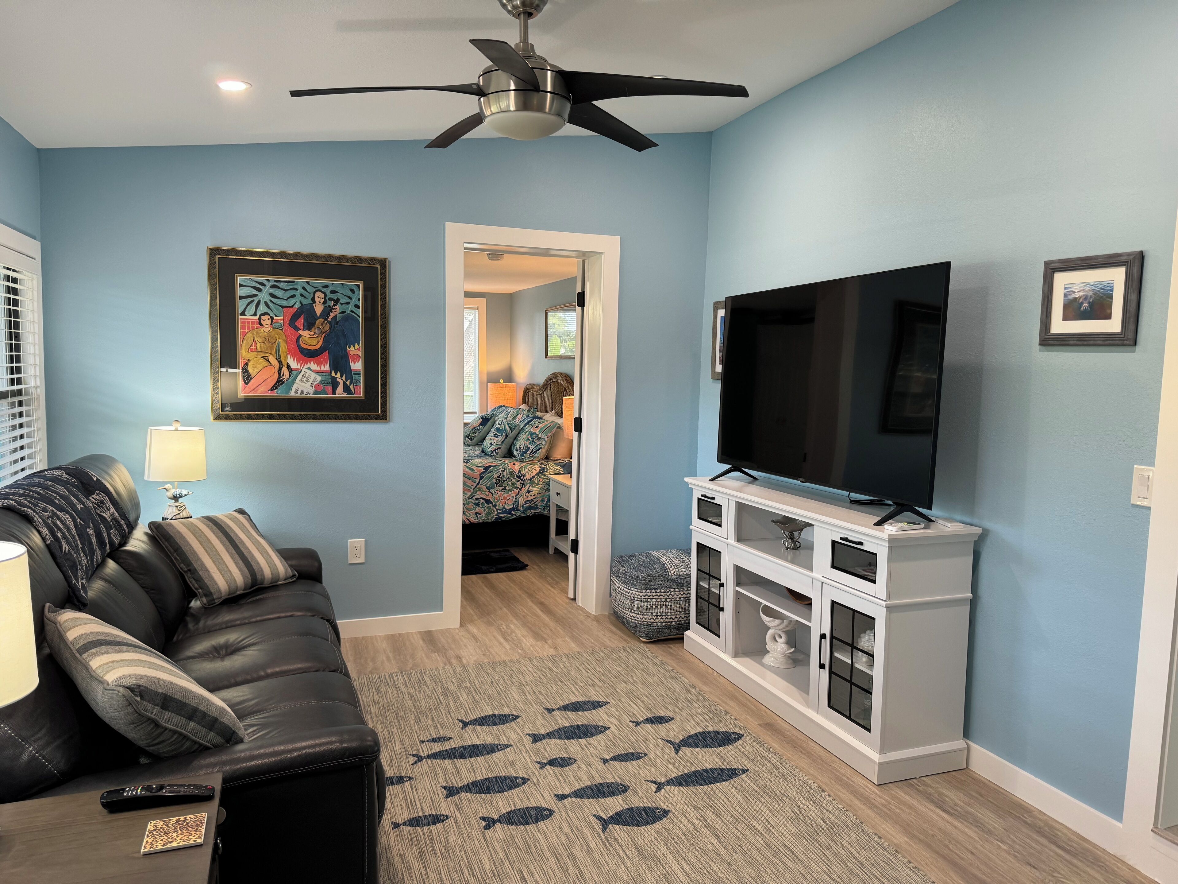 A living room with a black leather couch, a TV on a white stand, a ceiling fan, and a framed picture on the wall.