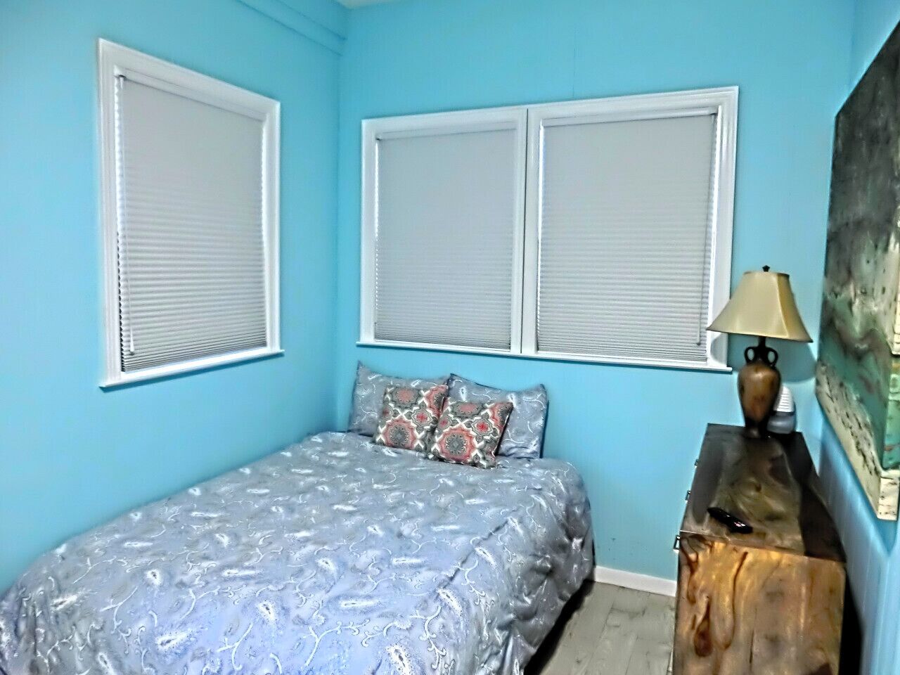 A bedroom with a bed, two windows with blinds, a bedside table with a lamp, and a painting on the wall.