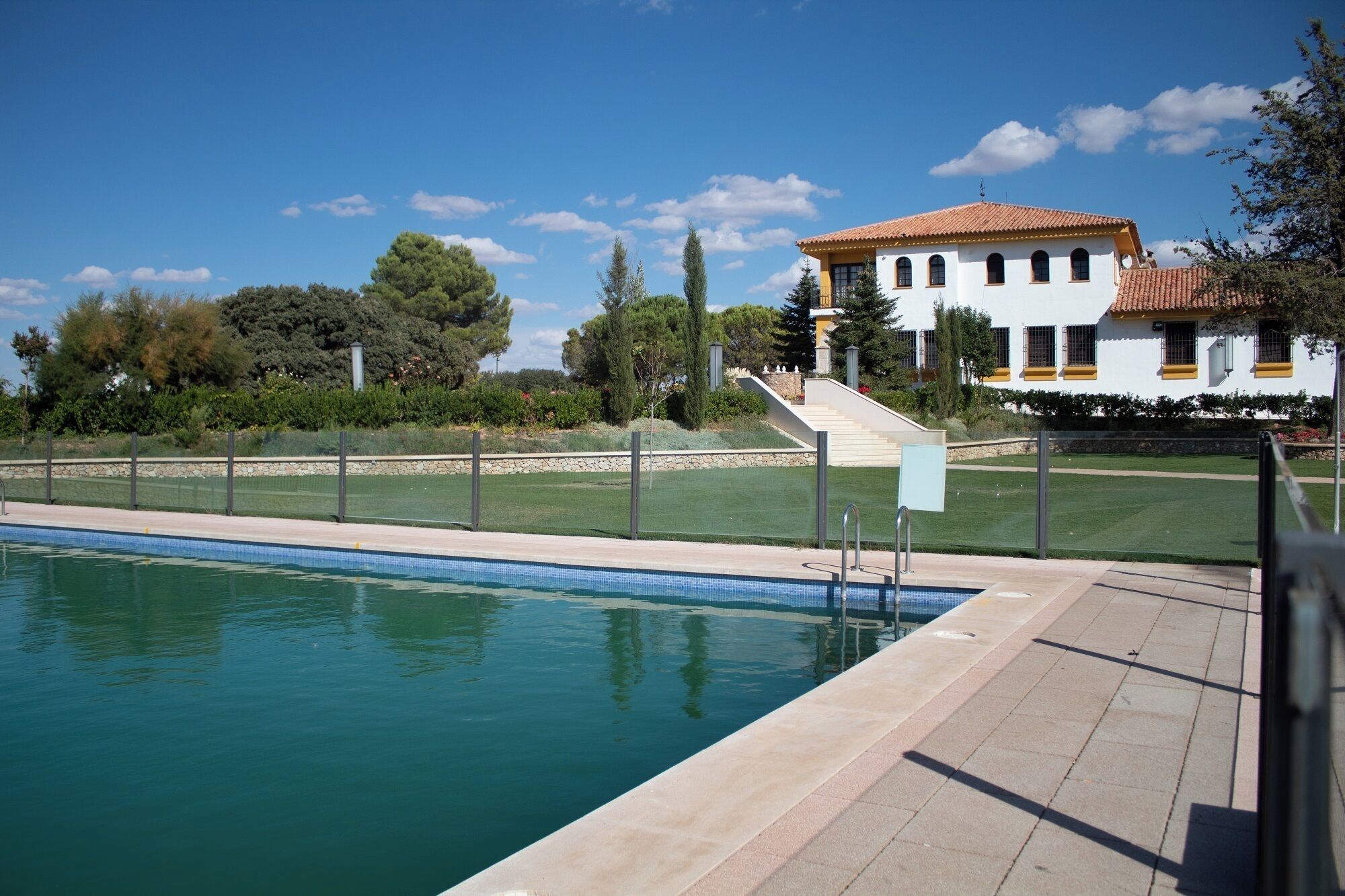 Piscina com superfície de água azul cristalina, ladeada por um deck pavimentado. Ao fundo, um edifício branco com telhado de telhas vermelhas e uma escadaria de pedra que leva a uma porta.